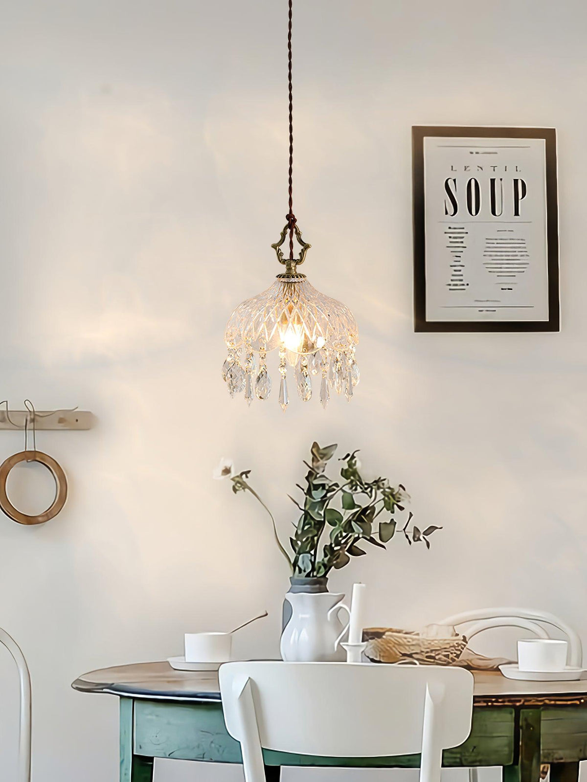 Clérachic crystal-drop pendant lit above a green-painted dining table with a vase of greenery, a white chair and a framed print on the wall.