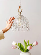 A hand reaching towards a lit Clérachic crystal-drop pendant, with pink tulips in a white vase on a pale wall below.