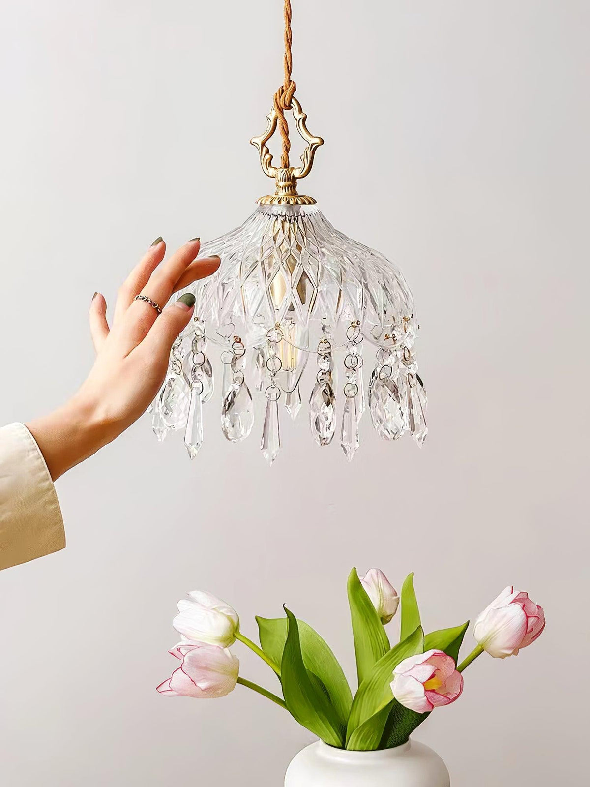 A hand reaching towards a lit Clérachic crystal-drop pendant, with pink tulips in a white vase on a pale wall below.