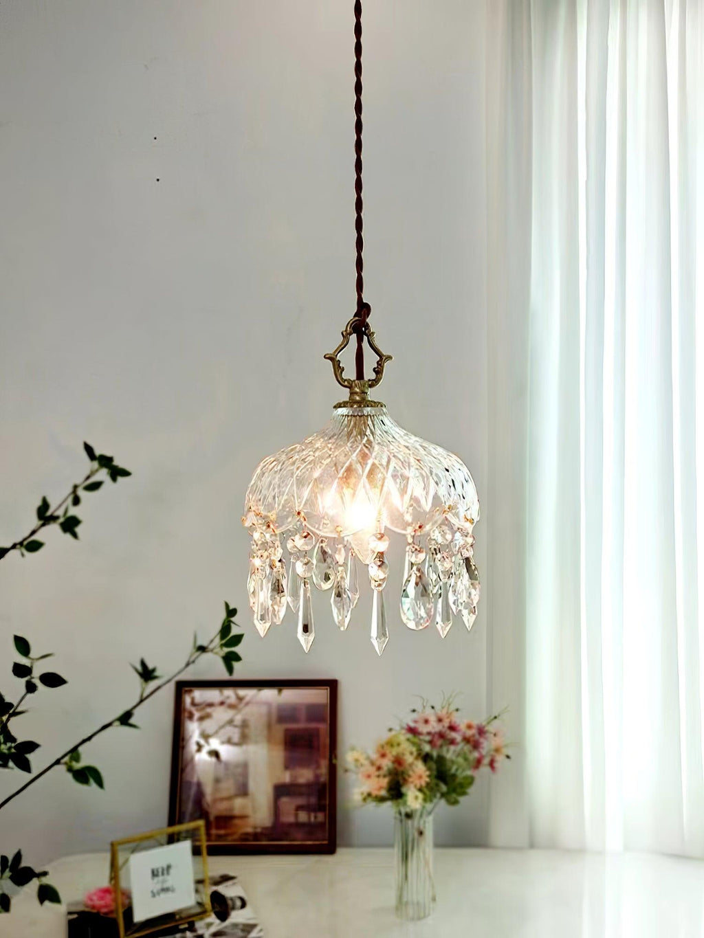 Clérachic crystal-drop pendant lit above a console with framed pictures and a vase of pink flowers, beside a sheer curtain.