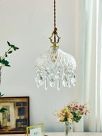 Clérachic crystal-drop pendant on a pale wall, with a framed picture and a vase of pink and peach flowers on a table below.