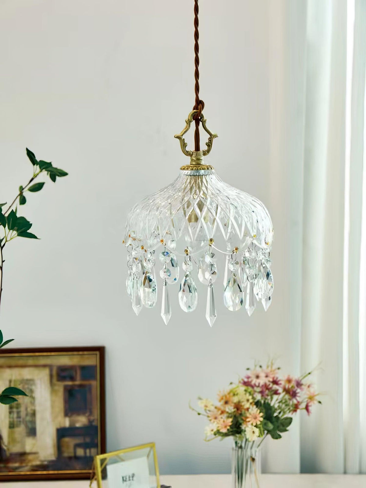 Clérachic crystal-drop pendant on a pale wall, with a framed picture and a vase of pink and peach flowers on a table below.