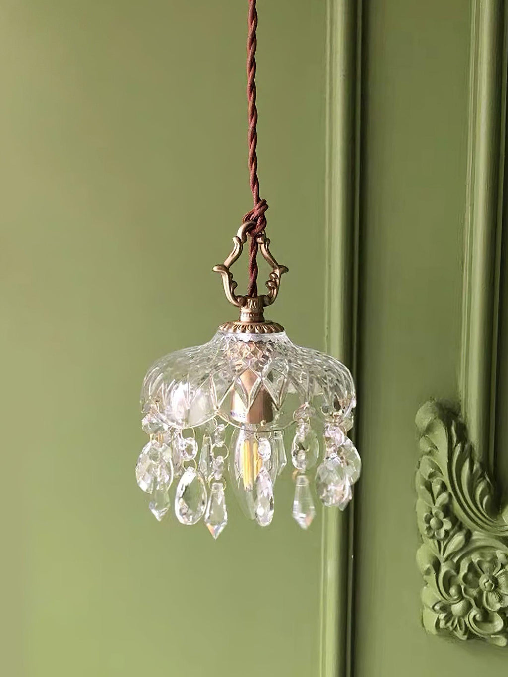 Clérachic crystal-drop pendant lit against a sage green panelled wall with an ornate carved moulding at the right.