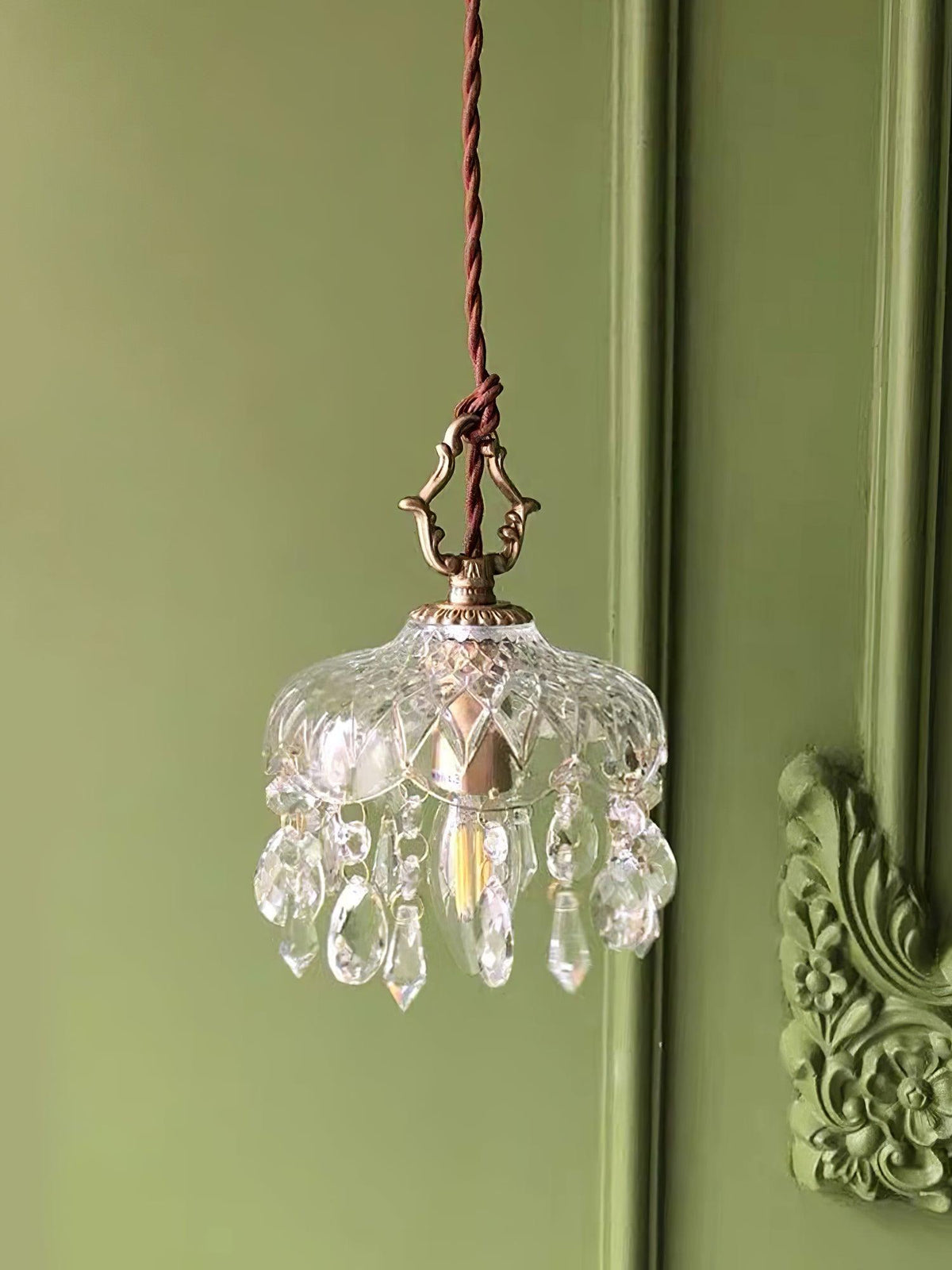 Clérachic crystal-drop pendant lit against a sage green panelled wall with an ornate carved moulding at the right.