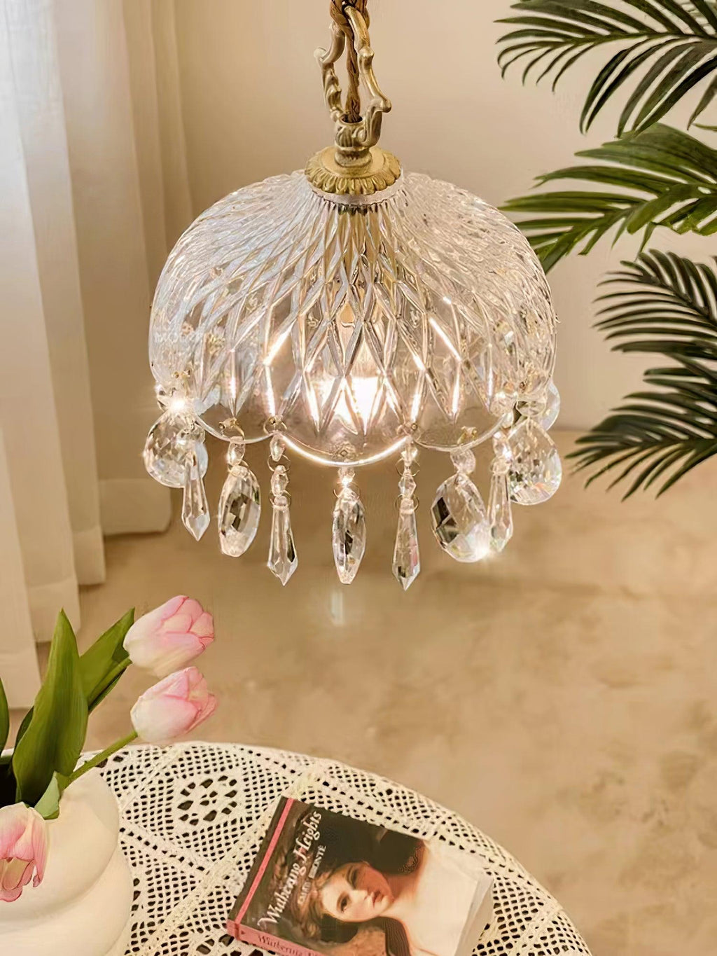 Clérachic cut-glass pendant glowing warmly above a marble-topped table with pink tulips and a lace cloth, with palm leaves behind.