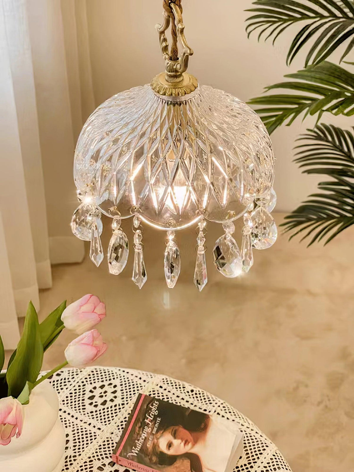 Clérachic cut-glass pendant glowing warmly above a marble-topped table with pink tulips and a lace cloth, with palm leaves behind.