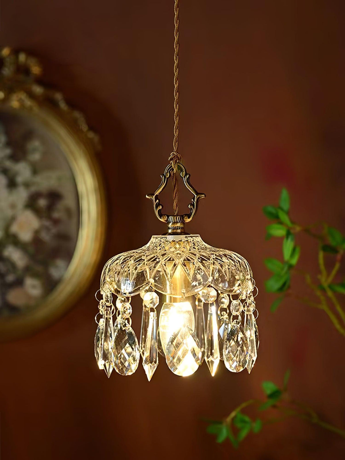 Clérachic crystal-drop pendant lit on a deep burgundy wall, with a gilt mirror frame at the left and green leaves at the right.