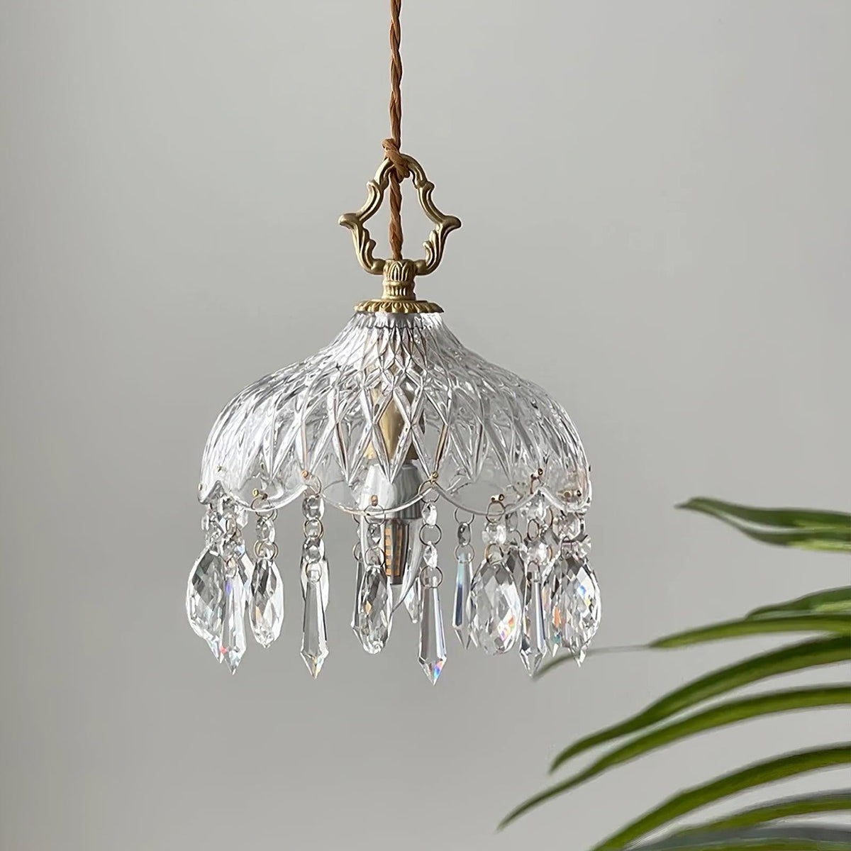 Clérachic crystal-drop pendant on a pale grey backdrop, with a diamond-cut glass bowl and a palm leaf at the right edge.