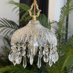 Close view of a Clérachic cut-glass pendant with its brass crown finial, set against dark palm leaves and a pale wall.