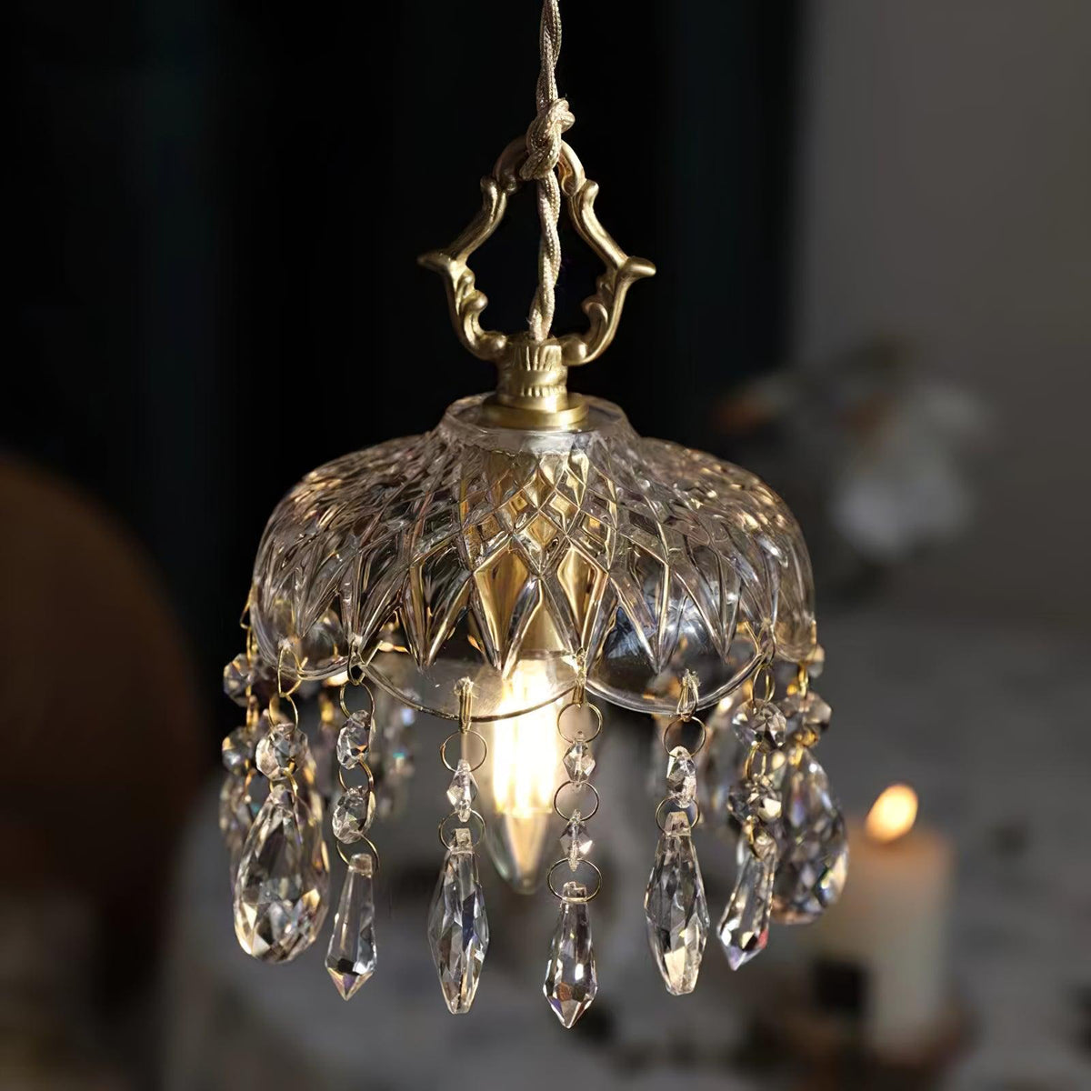 Clérachic crystal-drop pendant glowing on a dark backdrop, with a candle blurred behind and a brass finial above a smoke-tinted glass bowl.