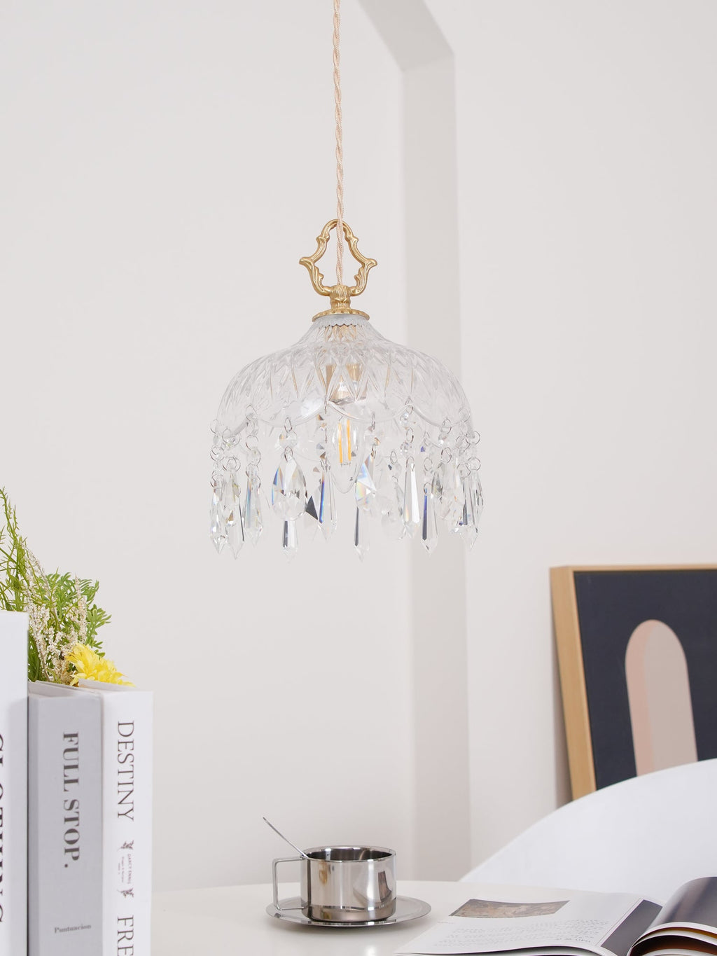 Clérachic crystal-drop pendant, pale and lit, hung against a white wall above a vase of flowers, books and an open magazine.