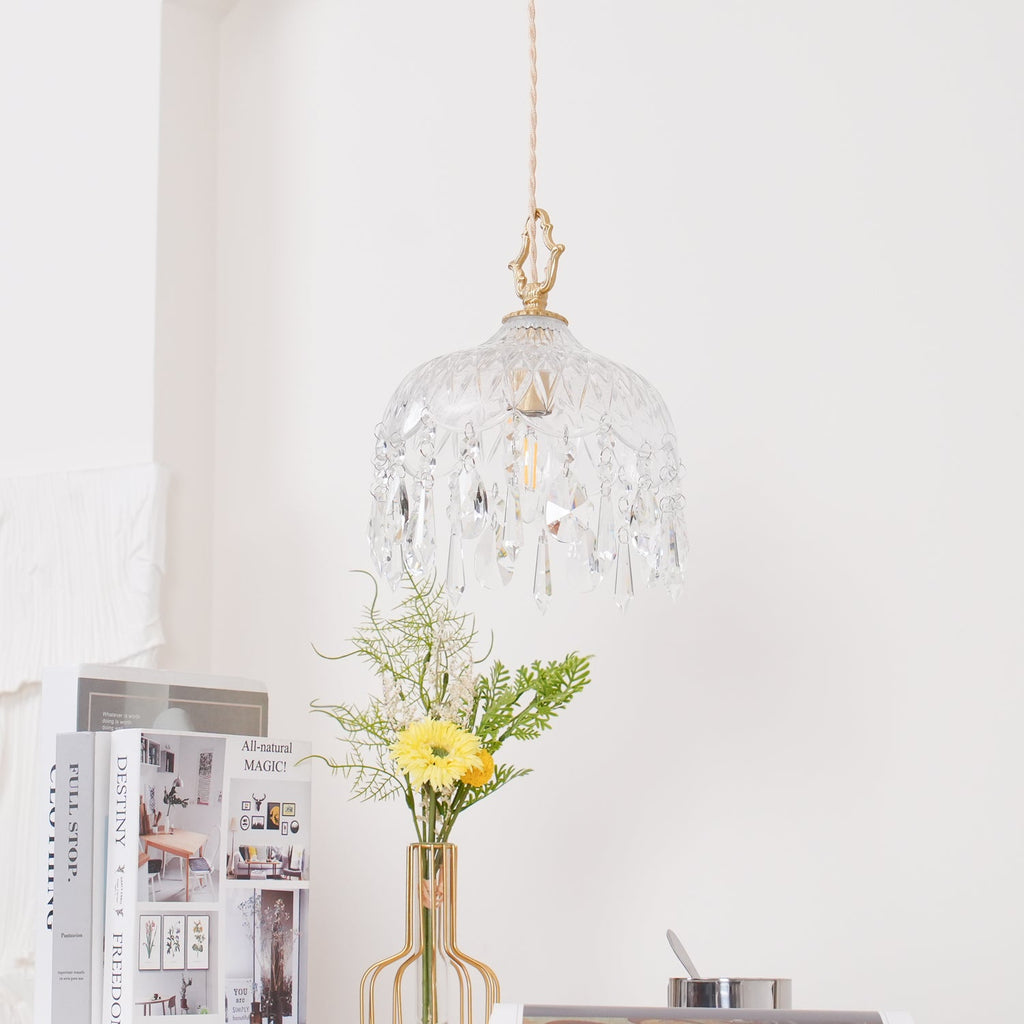 Clérachic crystal-drop pendant with a dark brass top, lit above a white table with a gold-framed vase of yellow flowers and books.
