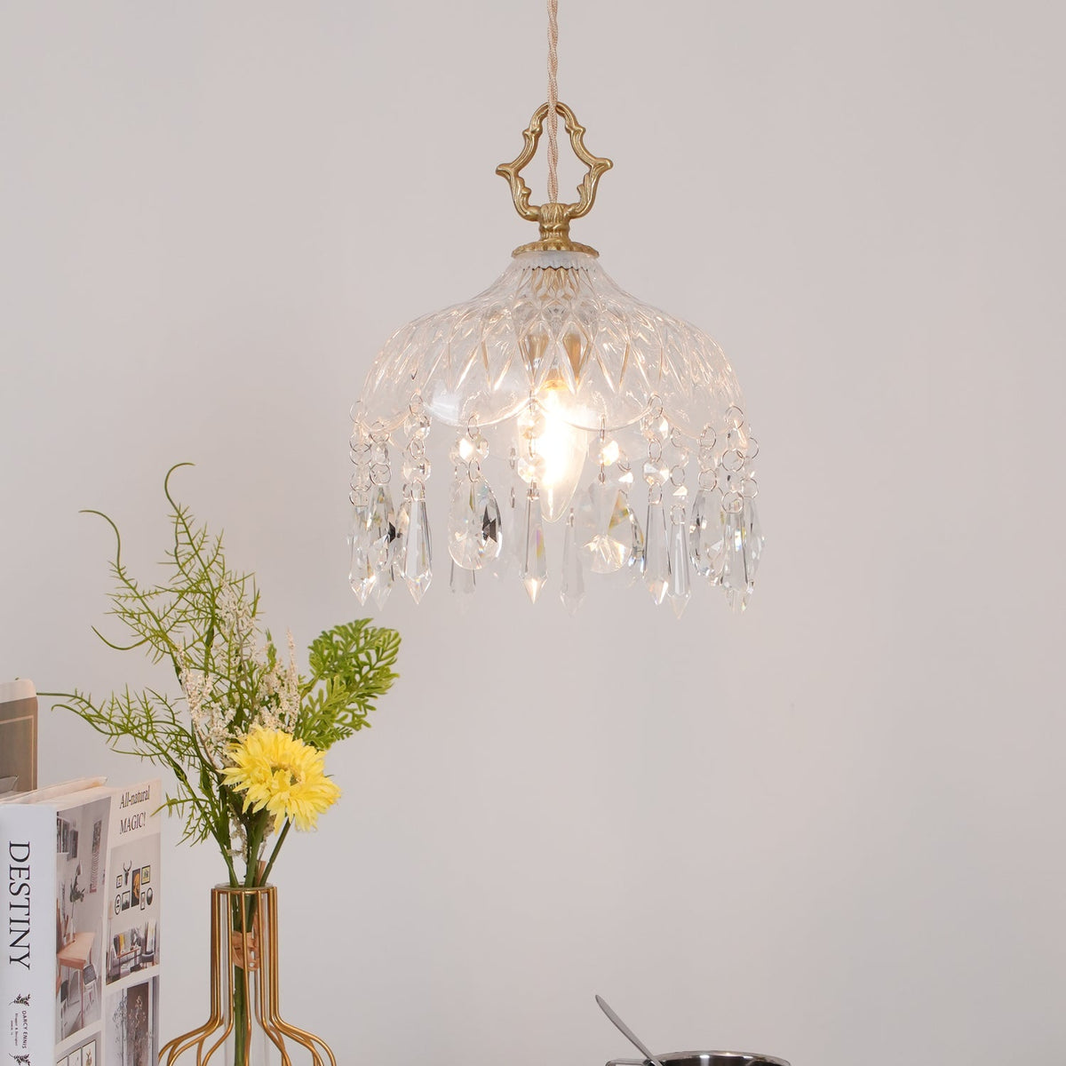 Clérachic crystal-drop pendant lit above a vase of yellow flowers on a white surface, with books and a steel cup at the lower left.