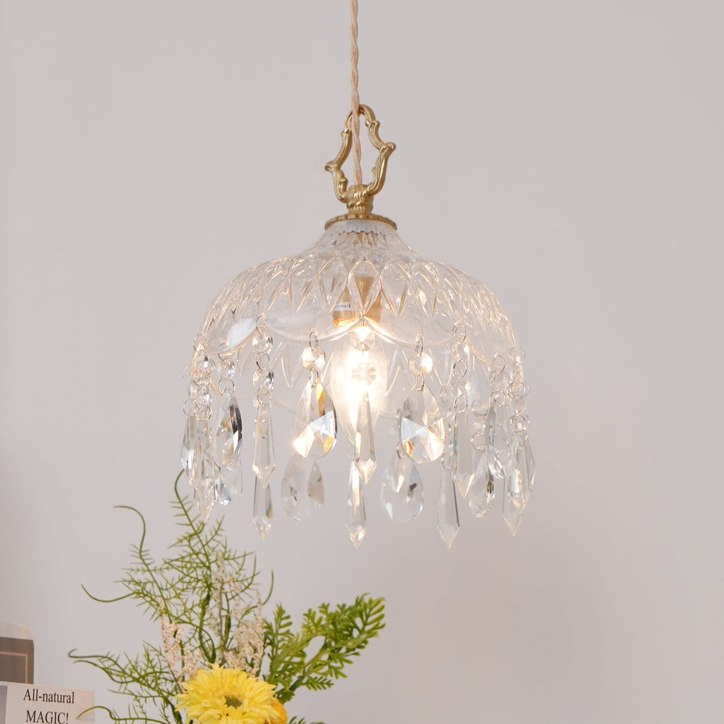 Clérachic crystal-drop pendant lit against a blush wall, with greenery and a yellow flower at the lower left.