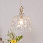 Clérachic crystal-drop pendant lit against a blush wall, with greenery and a yellow flower at the lower left.