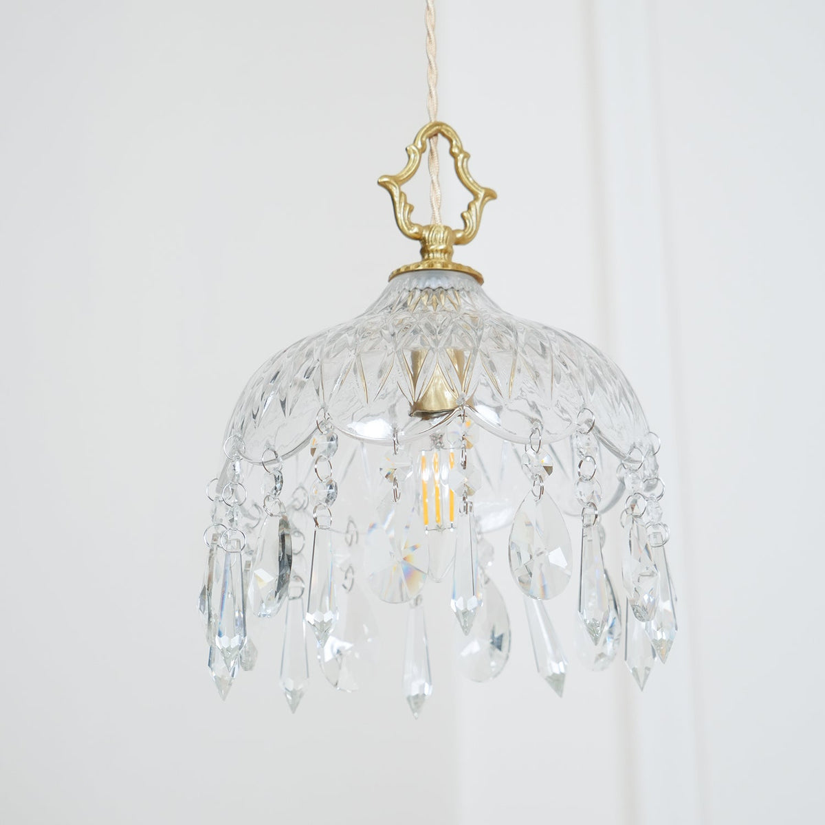 Clérachic crystal-drop pendant on a pale wall, with a diamond-cut glass bowl, a brass crown finial and rows of crystal drops.