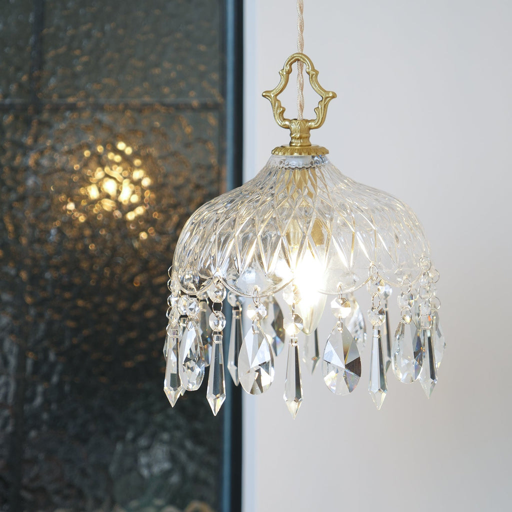 Larger view of a lit Clérachic crystal-drop pendant in front of dark patterned glass, with a brass finial and long crystal drops.