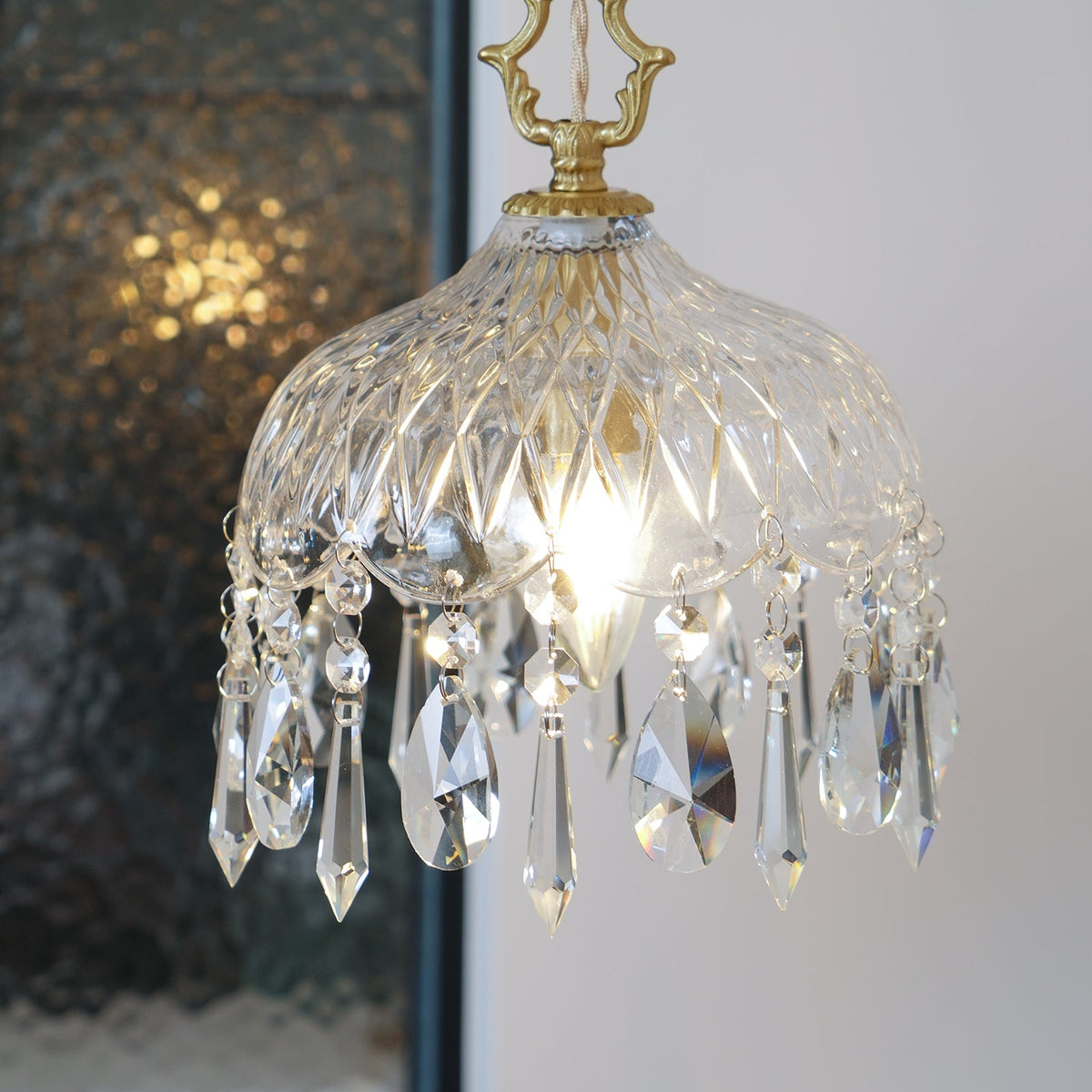 Clérachic crystal-drop pendant on a white backdrop, with a twisted dark cord and its candle bulb glowing through the glass.