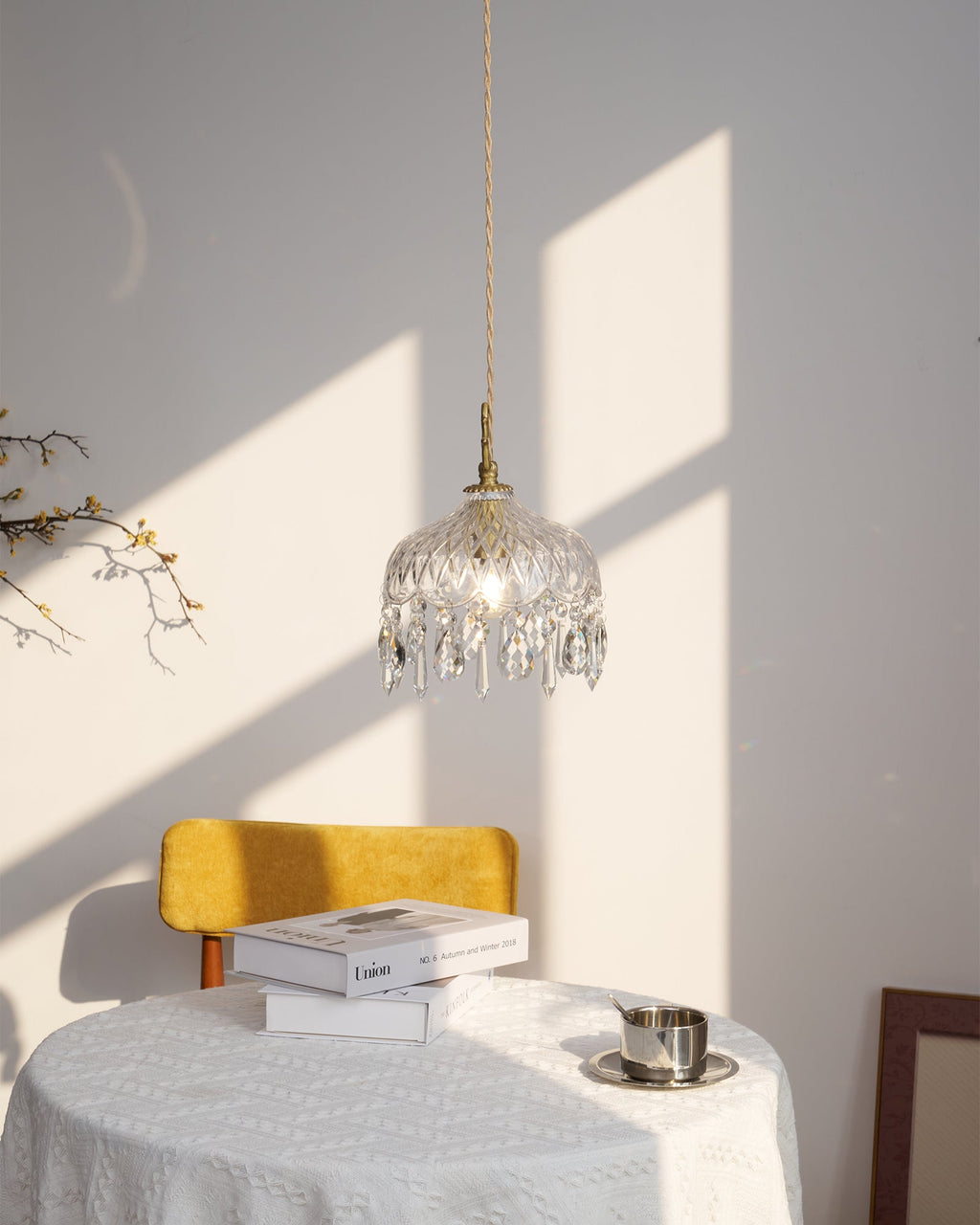 Clérachic crystal-drop pendant lit above a white table and a mustard chair, with books and a steel cup and a grey artwork behind.