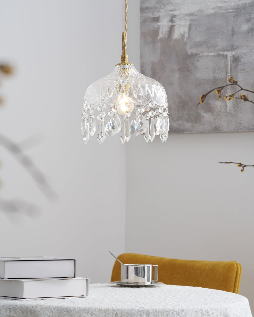 Clérachic crystal-drop pendant lit in a corner above a table with books and a steel cup, with a yellow chair at the right.