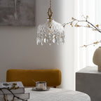 Clérachic crystal-drop pendant lit beside bare branches and a cream vase, with a mustard chair and a grey stone block at the right.