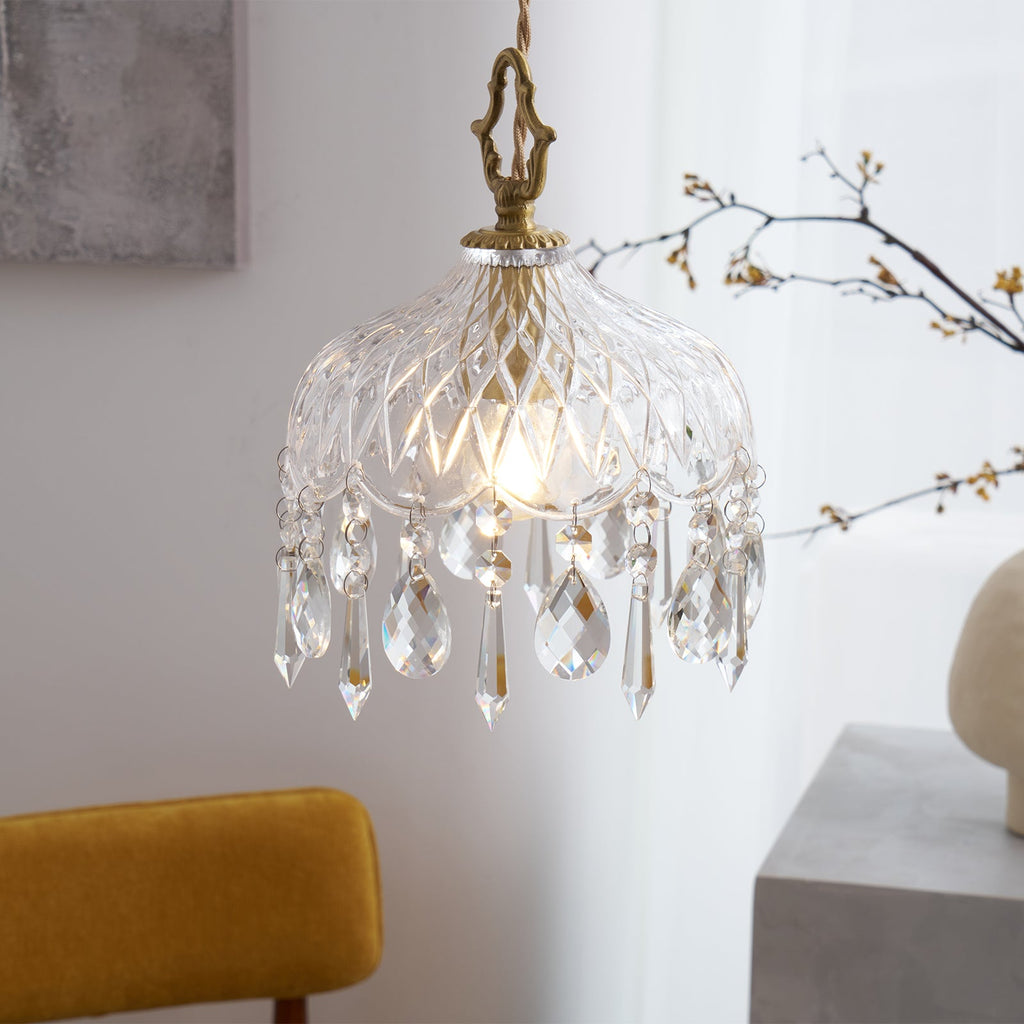 Closer view of a Clérachic crystal-drop pendant lit in a sunny corner, with a brass crown finial, a grey artwork and bare branches.