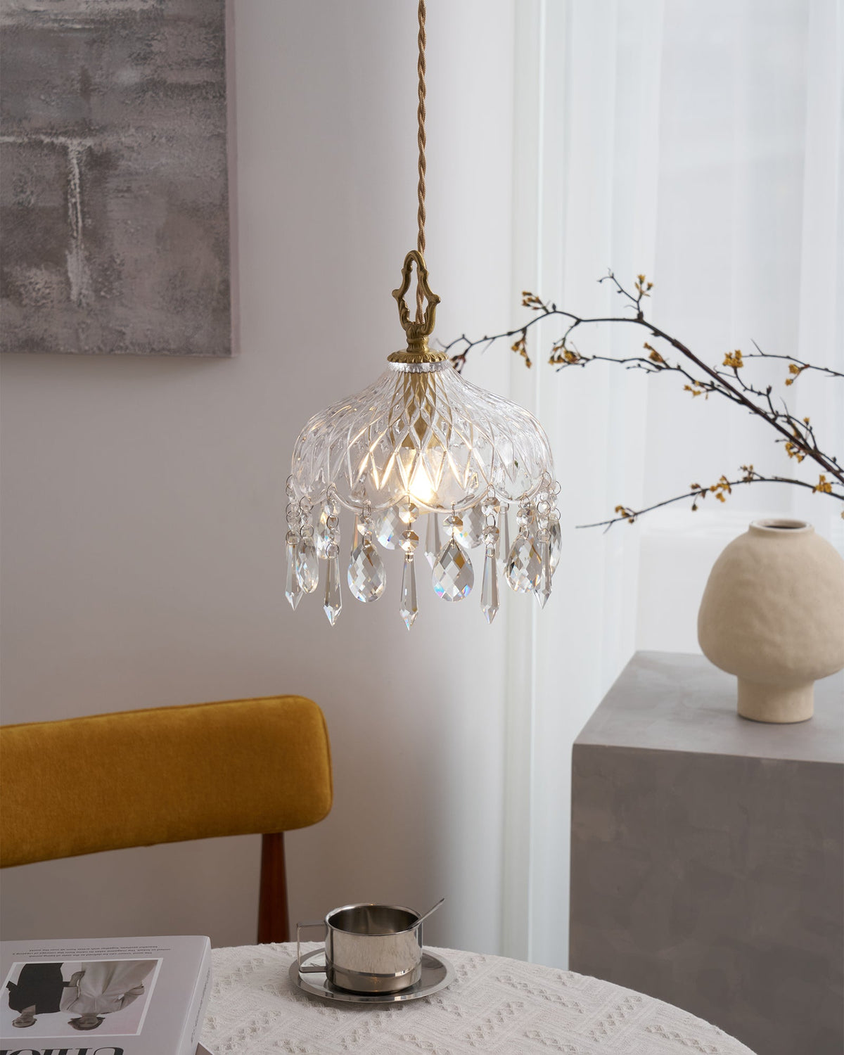 Clérachic crystal-drop pendant lit beside a grey artwork and a cream vase on a stone block, with a mustard chair in front.