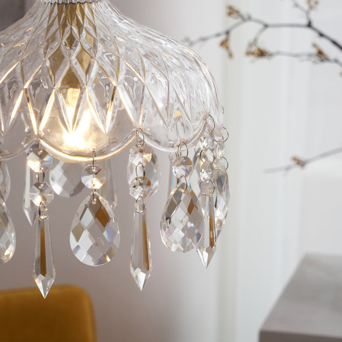 Close view of the diamond-cut glass and the crystal drops on this Clérachic pendant, with the bulb glowing and a branch behind.