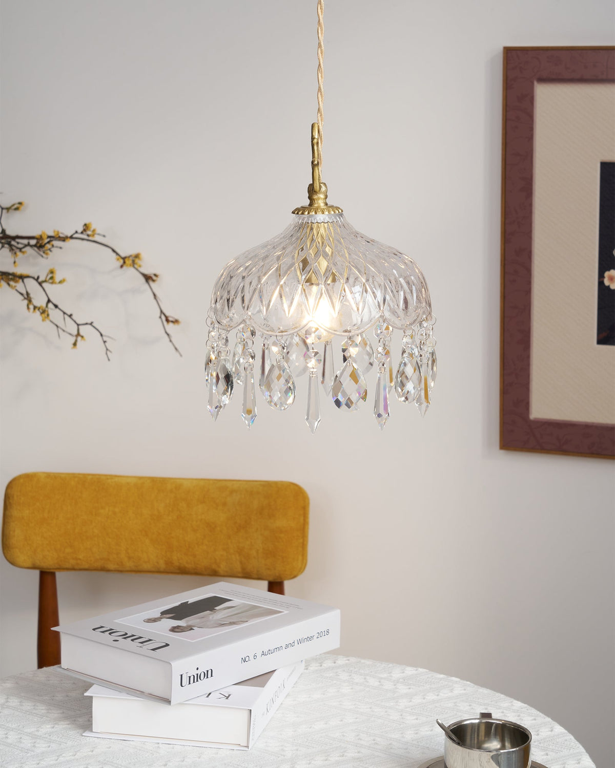 Clérachic crystal-drop pendant lit in a sunlit room, with a mustard chair, stacked books and a framed print with a dark mount.