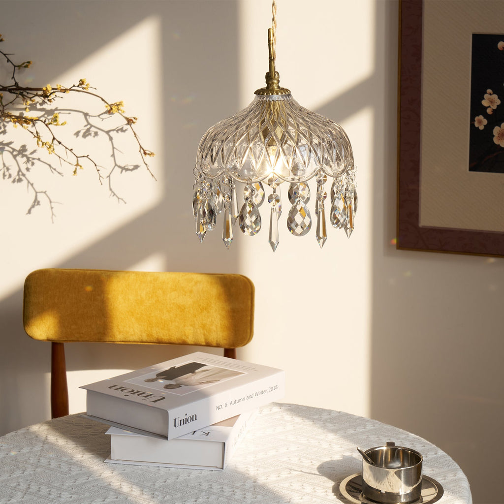 Clérachic crystal-drop pendant in strong sunlight and shadow, above a mustard chair and books, with a framed print at the right.