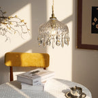 Clérachic crystal-drop pendant in strong sunlight and shadow, above a mustard chair and books, with a framed print at the right.