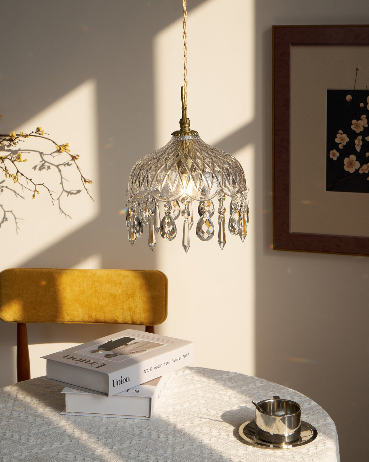 Clérachic crystal-drop pendant lit in warm light, above a mustard chair and a table with books and a steel cup.