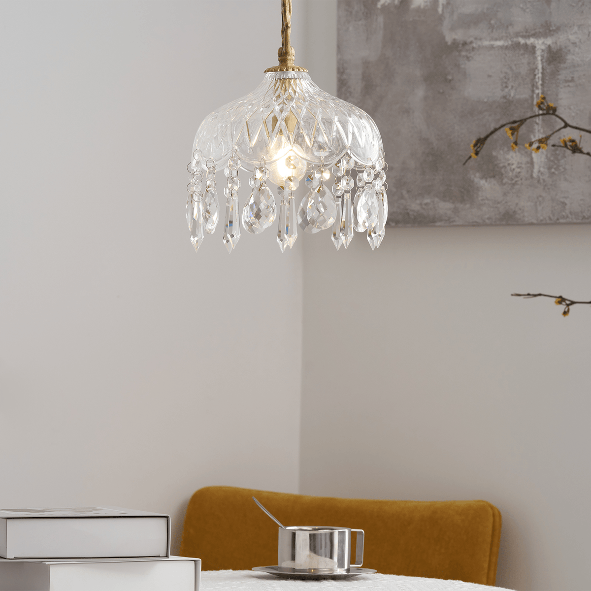 Clérachic crystal-drop pendant lit against a pale corner with a grey artwork, and a mustard chair and books at the bottom.