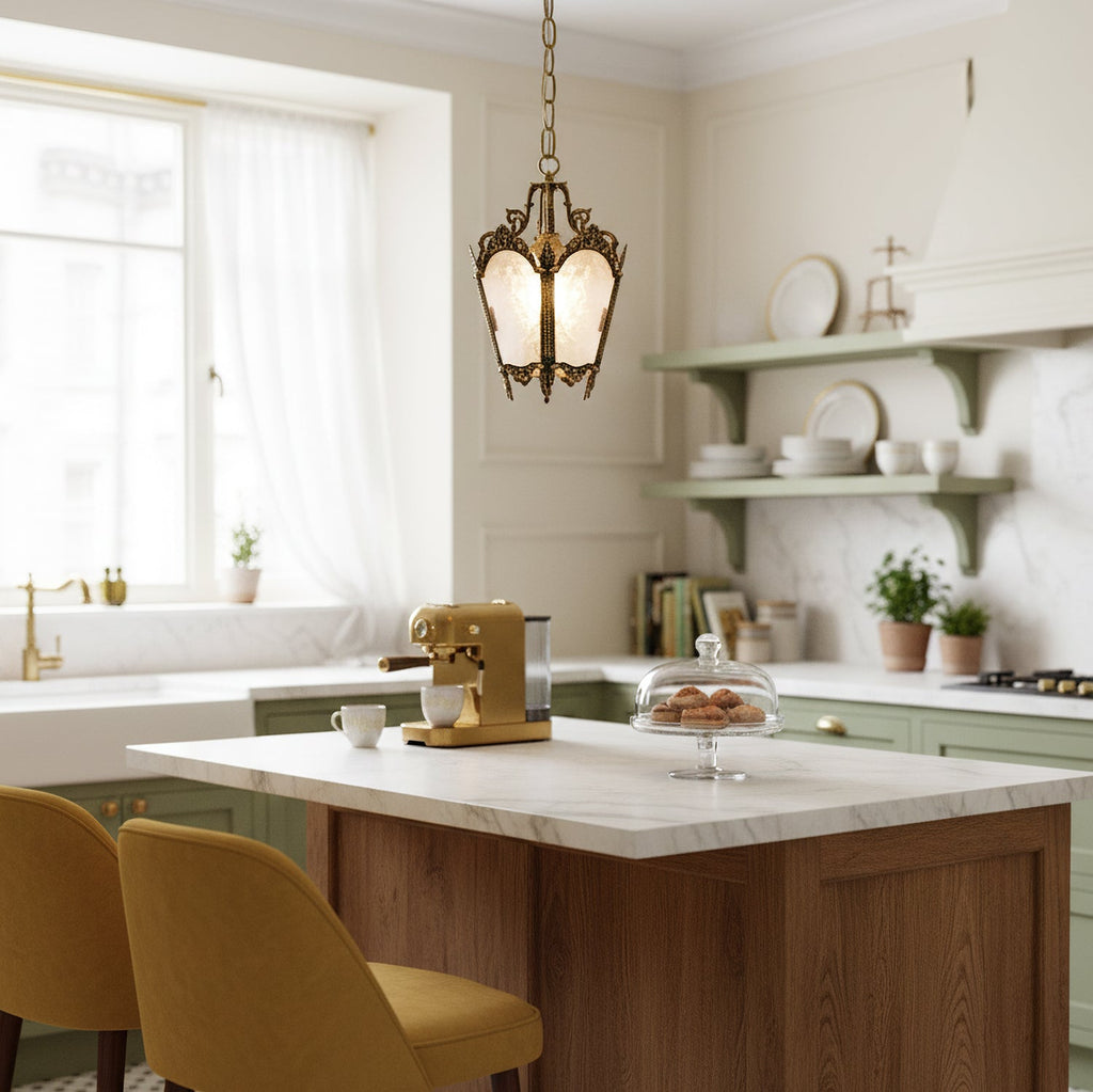 A brass Clérachic lantern pendant with frosted glass hangs above a marble island in a bright kitchen with mustard chairs and open shelves.
