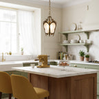 A brass Clérachic lantern pendant with frosted glass hangs above a marble island in a bright kitchen with mustard chairs and open shelves.