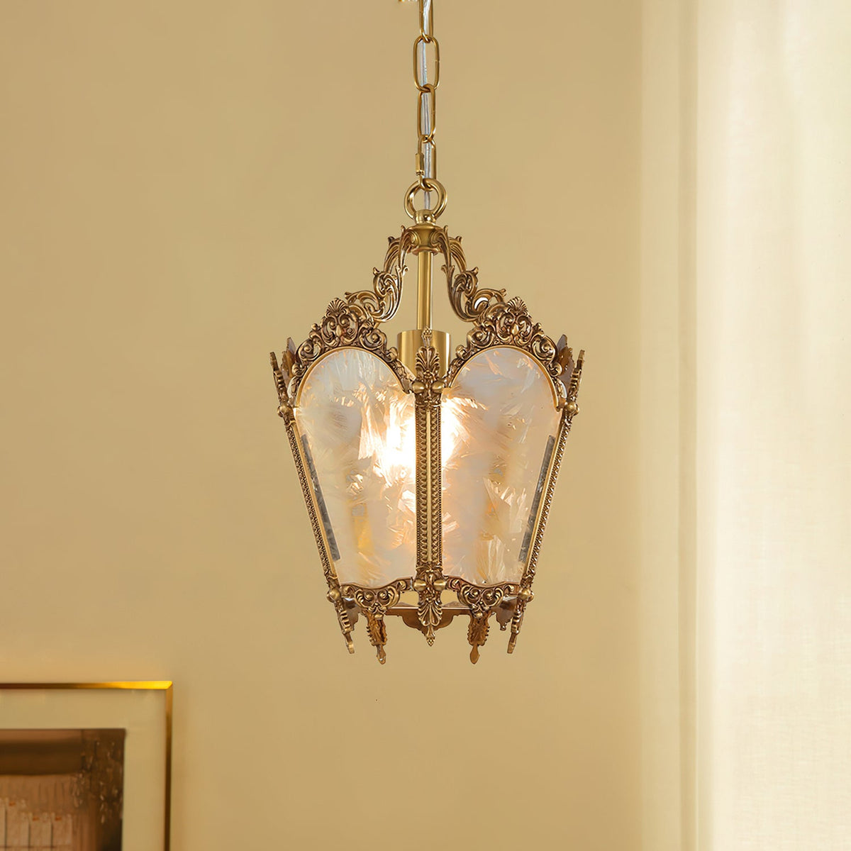A lit Clérachic lantern pendant with carved brass scrollwork hangs against a warm cream wall, beside a gilt picture frame.