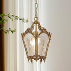 A Clérachic lantern pendant with a leafy branch beside it, its frosted glass panels glowing softly inside a patterned brass frame.