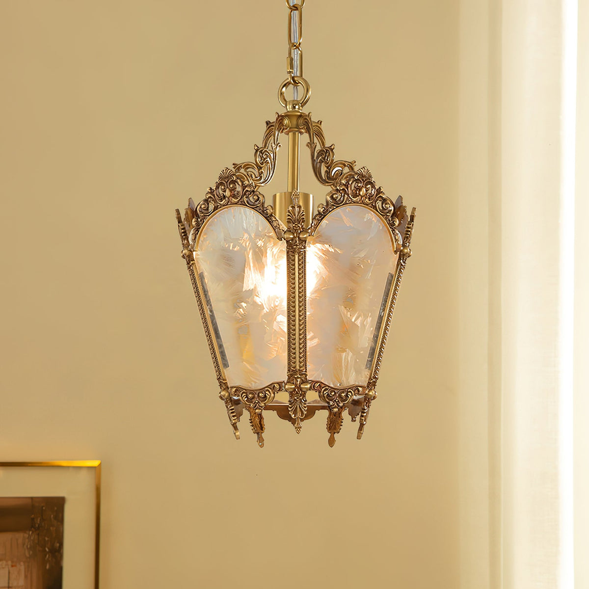 A lit Clérachic lantern pendant hangs in front of a cream curtain, with a gilt picture frame at the lower edge.