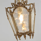 Close view of a lit Clérachic lantern pendant, with its frosted glass panels, patterned brass edging and small pendant drops at the base.