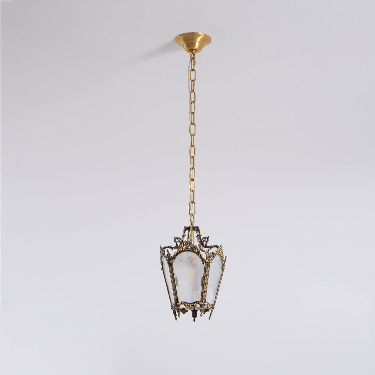 Seen from further away, a Clérachic lantern pendant hangs from a long chain and a brass canopy on a pale grey backdrop.