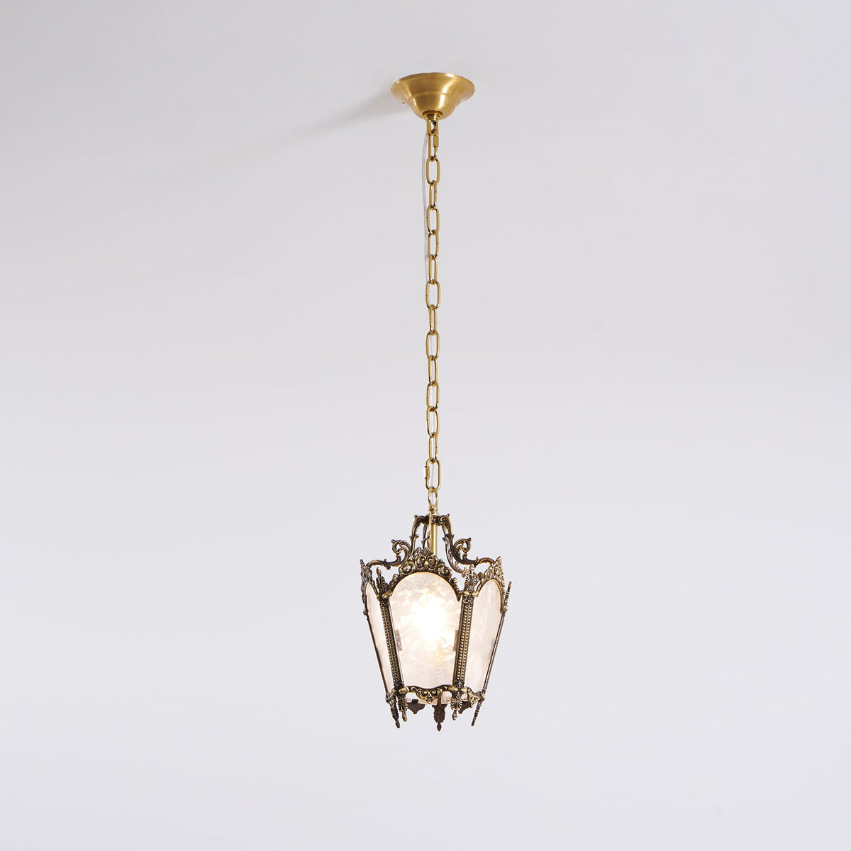 An unlit Clérachic lantern pendant with an aged brass frame hangs from a brass canopy against a plain pale wall.