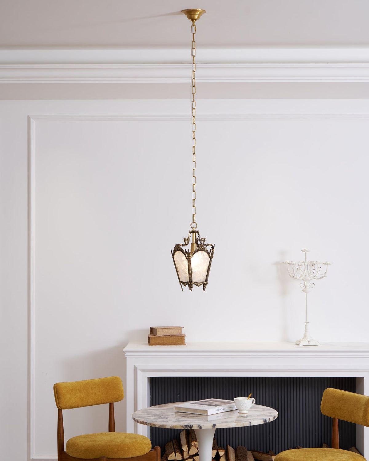 A Clérachic lantern pendant hangs from a long chain in a white room, above a marble table, a mantel and two mustard chairs.