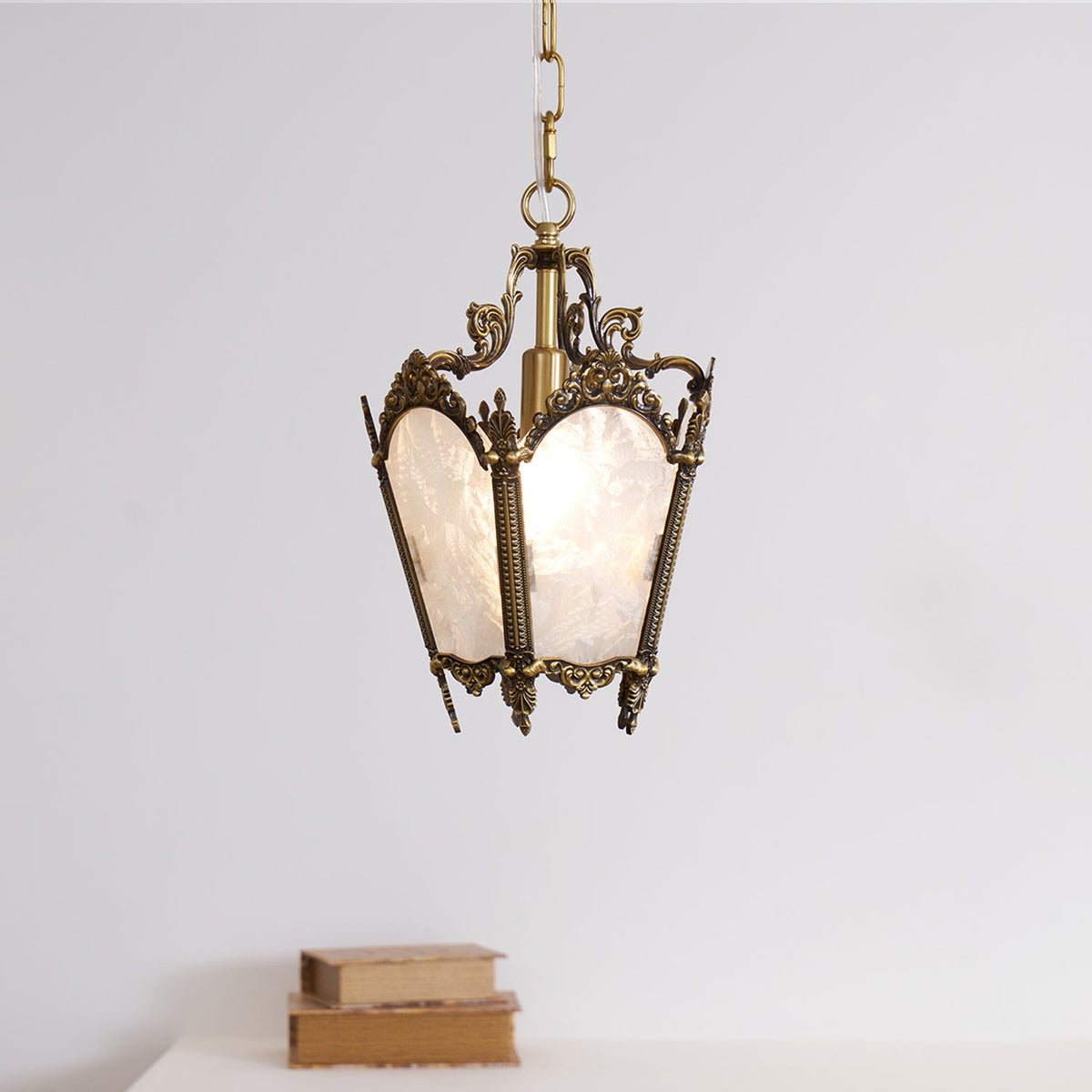 A Clérachic lantern pendant with an aged brass frame hangs on a chain above a white surface with two stacked books.