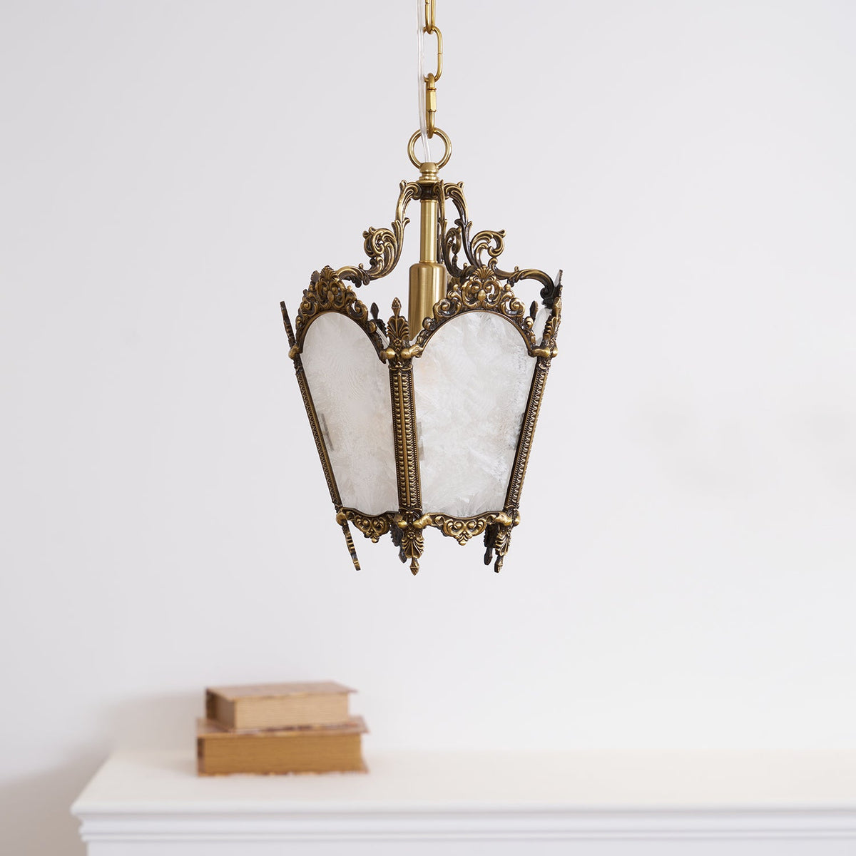 A Clérachic lantern pendant with white frosted panels and a darker brass frame hangs above a white mantel with two books.