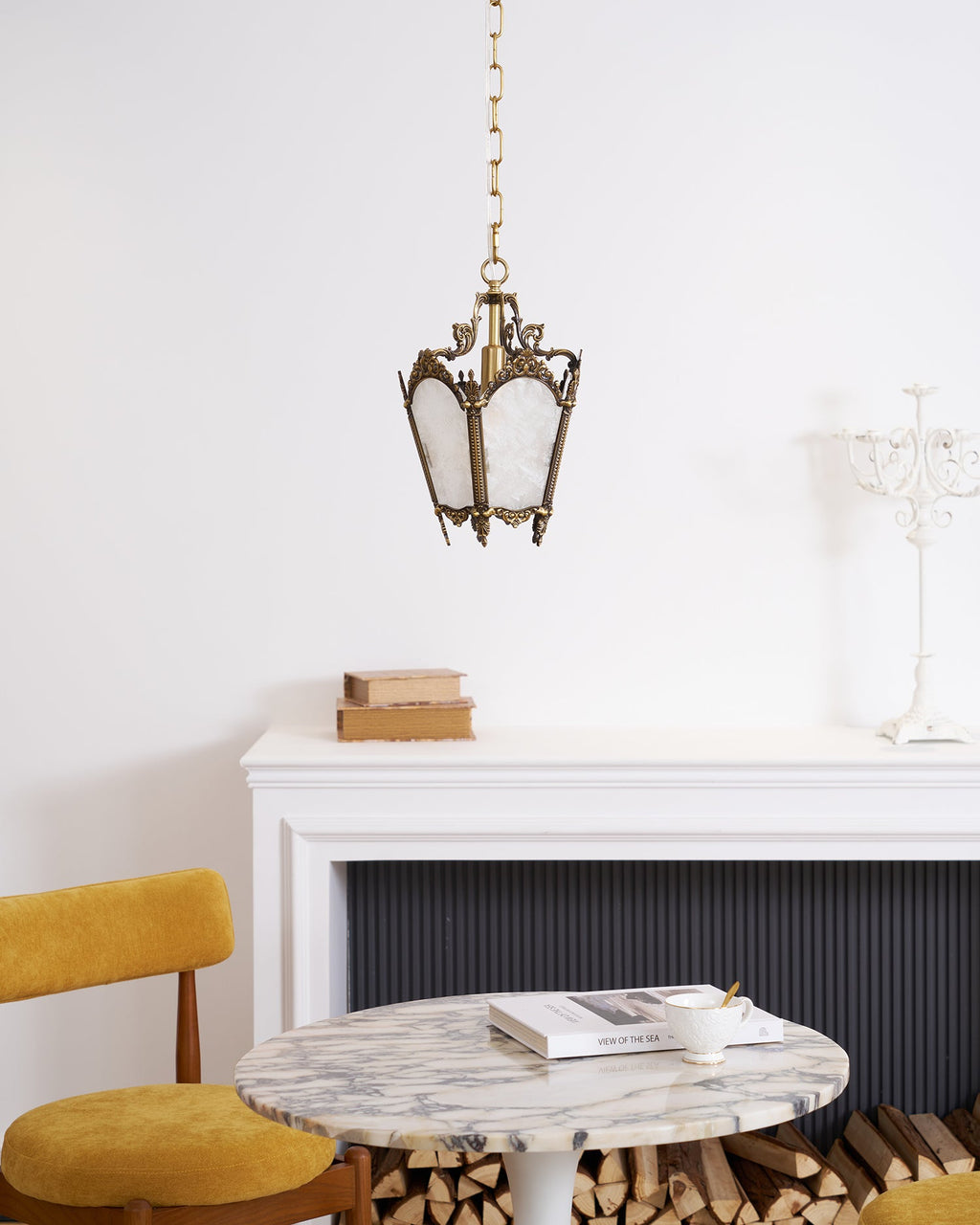 A Clérachic lantern pendant hangs in a white room above a mantel, a marble table and a mustard chair, with a white candelabra to one side.