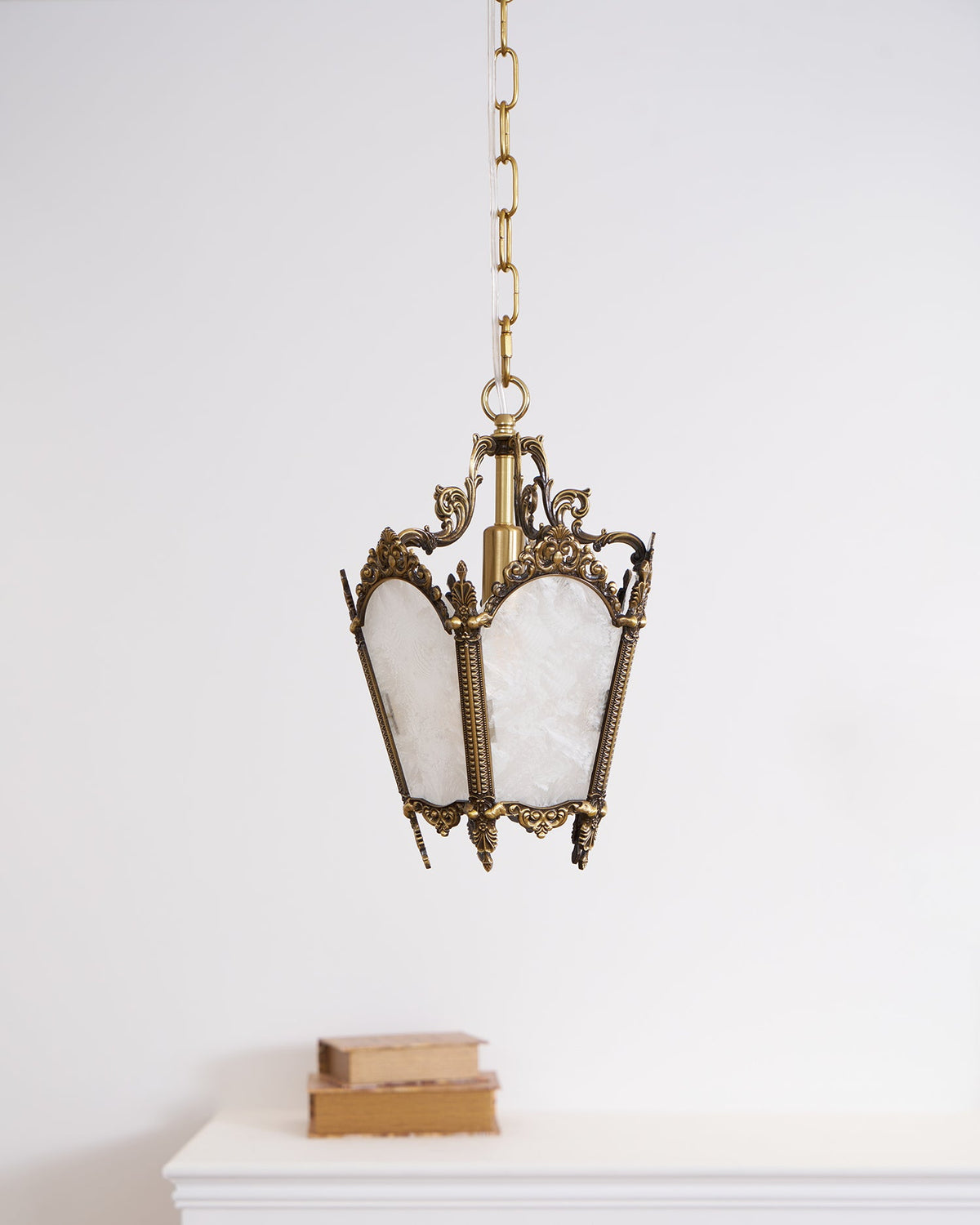 A Clérachic lantern pendant hangs on a chain above a white mantel with two books, against a plain white wall.