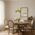 A green petal-edged Clérachic pendant hangs over a round wooden table with cane-backed chairs, beside a gold-framed mirror.
