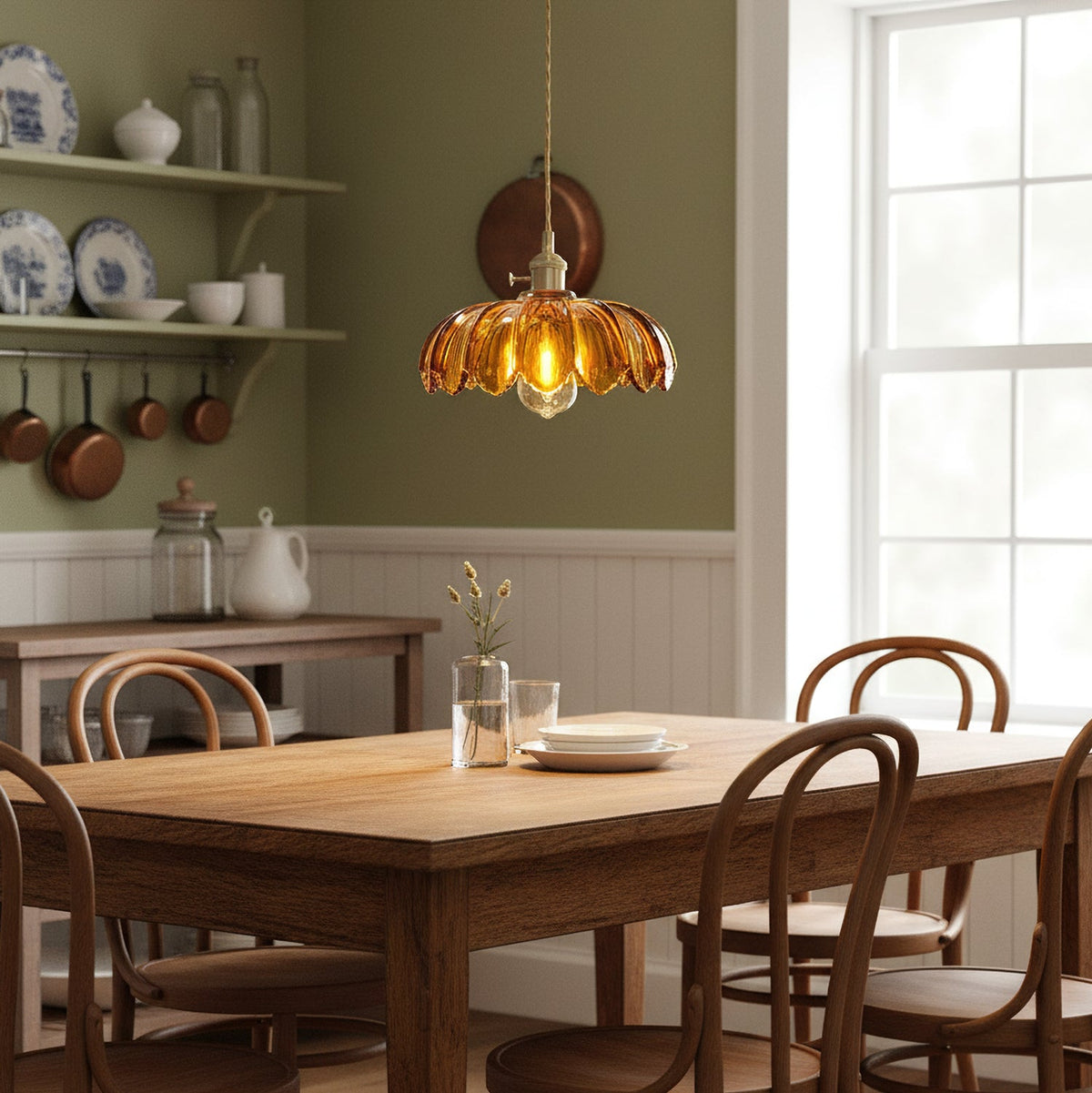Lit Clérachic pendant in amber glass above a long wooden kitchen table with bentwood chairs, in front of an olive green wall.