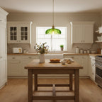 Small green Clérachic pendant glows over a window in a cream kitchen, beside a plain wooden island with a vase of dried flowers.