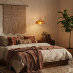 Amber glass Clérachic pendant lit above a bed with patterned red cushions and a macramé wall hanging, with a tall plant at the right.